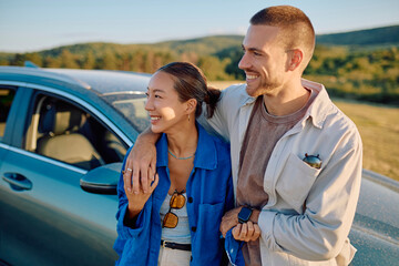 Young tourists smiling and hugging next to their electric car during a road trip in the countryside at sunset