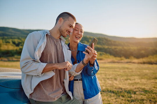 Young multi-ethnic couple smiling and using mobile phone during a road trip, leaning on their car in a field at sunset