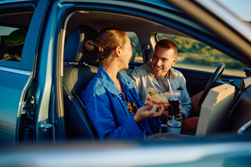 Happy couple eating pizza and drinking soda inside their car, enjoying a meal during a road trip