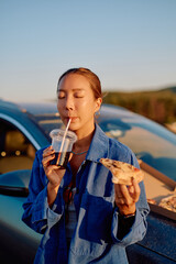 Asian woman drinking soda and eating pizza slice during road trip break near her car, enjoying...