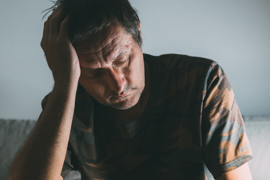 Pensive regretful middle-aged man sitting indoors in low light, deep in thought with hand on head