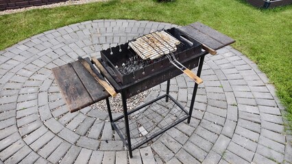 In the yard, next to the house and lawn, a metal brazier stands on a concrete tile. Under a metal...