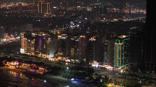 aerial view of Da Nang, Vietnam at night, showing tall buildings with sparkling lights, with My Khe Beach in the foreground. Busy streets. A modern coastal city.