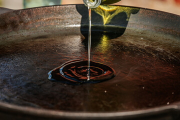 A stream of olive oil is being poured into a seasoned cast iron pan, creating a shiny pool in the...