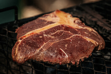Close-up of a thick raw T-bone steak sizzling on a grill with visible marbling and seasoning droplets in the air, capturing the moment of flavoring.