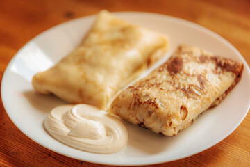 Two golden-brown stuffed crepes served on a white plate with a swirl of sour cream. The texture is soft and slightly crispy with a warm, homemade appeal.