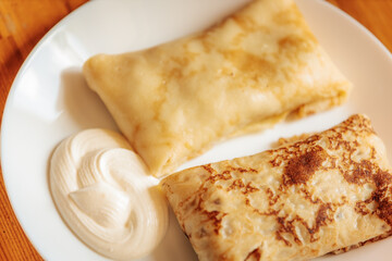 Two golden-brown stuffed crepes served on a white plate with a swirl of sour cream. The texture is soft and slightly crispy with a warm, homemade appeal.