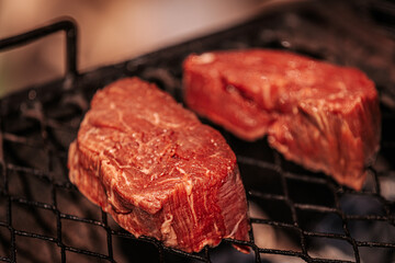 Two thick, raw beef steaks with visible marbling placed on a grill over open flames, ready for...