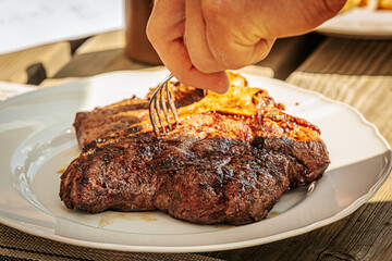 Close-up of a juicy grilled T-bone steak on a white plate, being sliced with a fork and knife. Cooked to perfection with visible grill marks and seared crust.