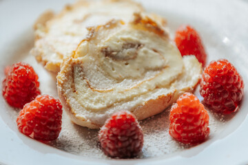 A close-up of a creamy roll cake slice topped with powdered sugar, surrounded by fresh raspberries on a white plate, creating a delicious and elegant dessert scene.