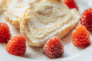 A close-up of a creamy roll cake slice topped with powdered sugar, surrounded by fresh raspberries on a white plate, creating a delicious and elegant dessert scene.