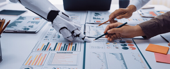 A futuristic scene depicting a collaboration between a human hand and a robotic arm over business reports, showcasing data analysis in a modern office environment.Noogenesis