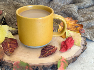 Warm coffee in a mustard yellow mug sits on a wooden slab surrounded by fall leaves and a knit...