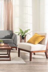 Mid-century modern inspired indoor seating area with light beige armchair and mustard cushion, charcoal grey sofa, dark wood coffee table, tropical plant, and natural morning sunlight ambiance.