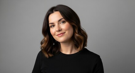 Fototapeta premium Smiling woman with dark hair and brown highlights poses against a neutral gray backdrop, radiating a friendly and approachable demeanor.