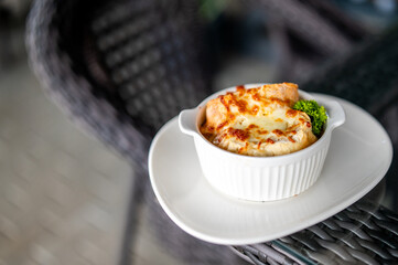 French onion soup in a white ceramic ramekin, topped with melted cheese and parsley, served on a white plate over wicker surface, warm and hearty comfort food, close-up view.