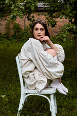 A thoughtful young woman with a headband and linen dress sits on a white chair in a green garden,...