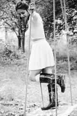 A black and white photo of a woman standing on a swing with a cheerful expression, wearing a white...