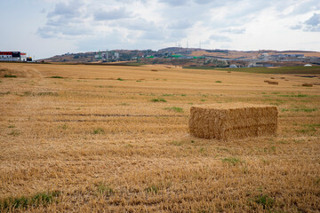 A vast rural landscape featuring a large, rectangular bale of hay in the foreground. The hay bale is golden brown and neatly stacked.