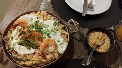A delicious plate of creamy shrimp risotto served in a rustic copper pan, garnished with fresh chives and pepper. A side of farofa and a plate with cutlery on a dark wooden table in a restaurant.