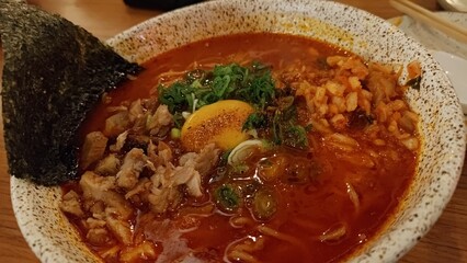 Close-up of a delicious bowl of spicy Japanese ramen noodle soup with pork, kimchi, scallions, nori seaweed, and an egg yolk. Traditional Asian cuisine served in a rustic ceramic bowl.