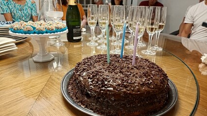 A vibrant birthday party table with a rich chocolate cake topped with sprinkles and candles. Flutes of sparkling wine are ready for a toast, with Brazilian sweets (brigadeiros) and guests in the backg
