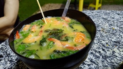Close-up of a Tacacá, a traditional, savory shrimp soup served hot in a rustic dark bowl on a granite table. This delicious seafood dish features a flavorful yellow broth with fresh herbs and spices.