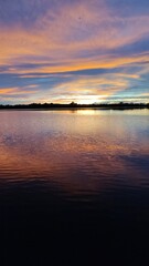 Breathtaking sunset over Amazon Jungle River, with vibrant orange and purple clouds reflecting on the calm water surface. A silhouetted forest lines the distant horizon under a dramatic, colorful sky.