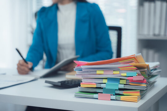 A businesswoman is working with a large pile of paperwork, searching for unfinished documents, information on piles of documents on her desk, and checking financial documents amidst her busy workload.