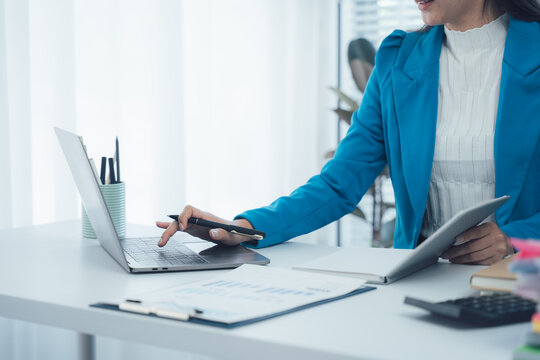 Focused Workflow: A professional woman in a stylish blue blazer intently works at her desk, seamlessly navigating a laptop and tablet, the scene bathed in natural light, emphasizing productivity.