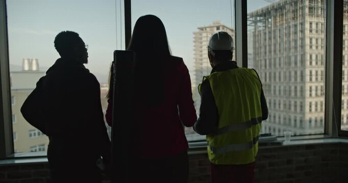A multinational business team discusses construction plans in a high-rise office building. The team examines blueprints in preparation to proceed.