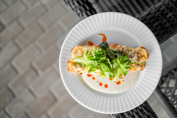 Stuffed crepe topped with greens, white and red sauces, served on a white plate. Gourmet presentation on textured background. Perfect for culinary and dining themes.