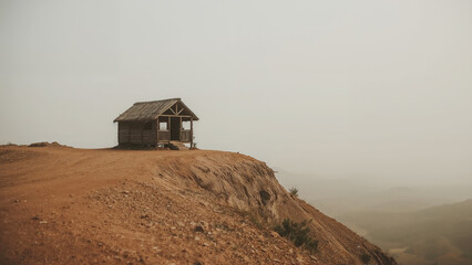 Small thatched roof hut perched on a dusty cliff edge overlooking a misty valley Keywords: hut, cabin, shack, dwelling, shelter, remote, isolated