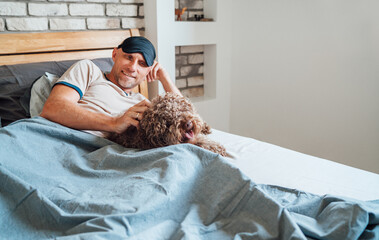 Maltipoo dog lying in bed with owner waking UP wearing black sleep eye mask after daytime nap on unfocused background. Funny pets and healthy lifestyle concept image.