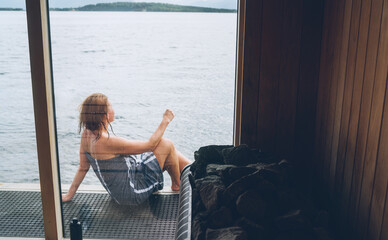 Relaxing Woman wrapped towel sitting on wooden Scandinavian floating sauna deck with huge wide...