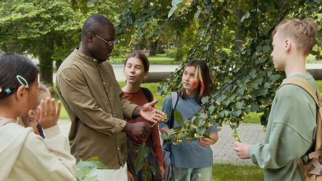 African American young male botanist with magnifying glass speaking with group of diverse school children while standing together under branches of linden tree during outdoor lesson