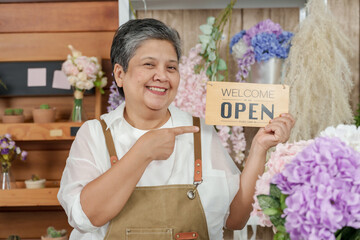 Senior asian female florist proudly pointing at wooden open sign while standing among flower...