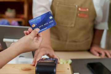 closeup hand holding credit card over wireless payment machine in small flower business shop operated by asian senior female florist processing transaction at wooden counter near tablet
