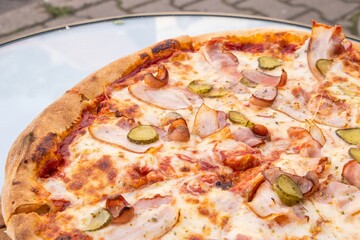 Delicious pizza topped with ham, pickles, and cheese, presented on a glass table, showcasing a mouthwatering culinary creation with vibrant colors and textures
