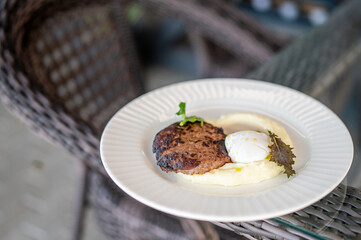 Grilled meat patty with mashed potatoes and poached egg, garnished with greens, served on a white plate outdoors, presented in a rustic wicker setting.