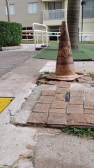 Worn and dirty traffic cone marking a hazardous pothole on a broken concrete and paver sidewalk....