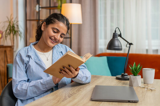 Indian young woman reading book, turning pages smiling enjoying literature, taking a rest sits at table desk. Portrait of peaceful cheerful girl relaxing at home apartment living room. Lifestyle.