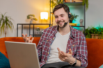 Happy young business man helpline in plaid shirt talking to web camera headset making an online video conference call in distance. Caucasian freelancer guy using laptop on couch in living room at home