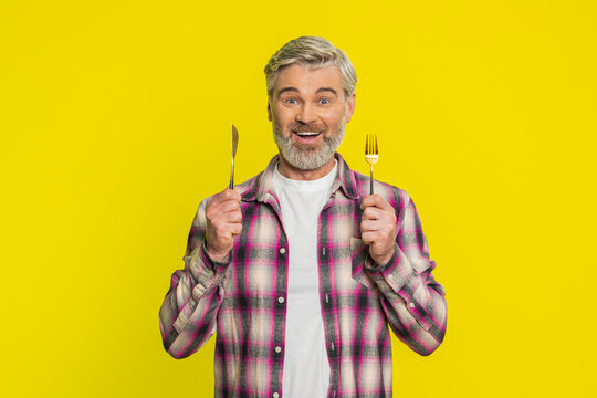 Ready to eat. Hungry Caucasian middle-aged man waiting for serving dinner dishes with with restlessness holding cutlery fork knife, will appreciate delicious restaurant meal on yellow background