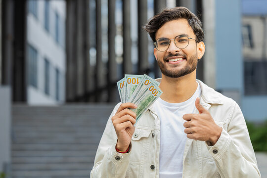 Happy Indian man counting money cash holding dollars banknotes, bills budget count salary profit reward bribe outdoors. Successful rich Arabian Hindu guy showing thumbs up like in downtown city street - Powered by Adobe