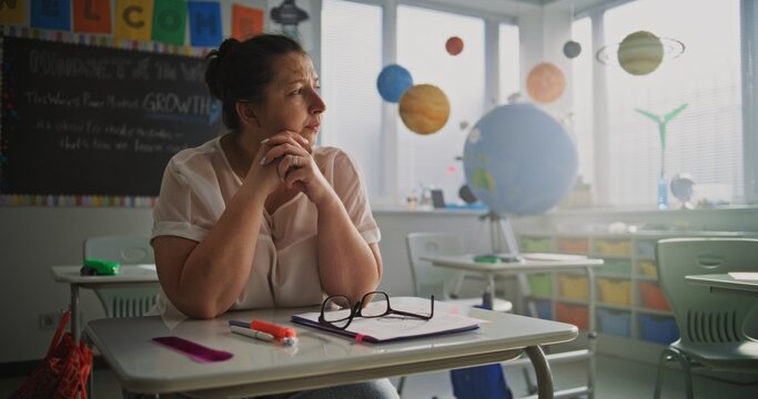 Tired Female Teacher Sitting Alone at the Desk in Empty Classroom, Relaxing After Class. Woman Feeling Stress, Burnout and Mental Exhaustion in Educational Environment, Working in Elementary School. - Powered by Adobe