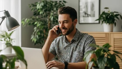 A young man smiling while talking on the phone in a modern home office filled with greenery working on a laptop with framed pictures on the wall and natural light streaming in - Powered by Adobe