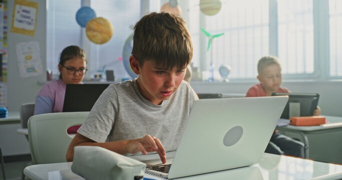 Elementary School Boy Sitting at Desk, Using Laptop in Classroom. Team of Smart Diverse Kids Working on Their Laptops and Tablet Computers, Studying Computer Science, Basic Programming and Coding.