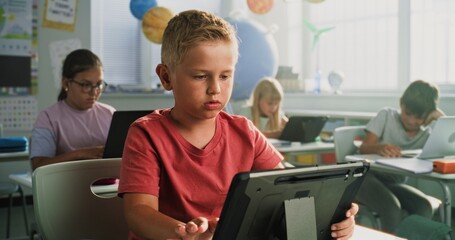 Elementary School Boy Sitting at Desk, Using Digital Tablet in Classroom. Team of Smart Diverse...