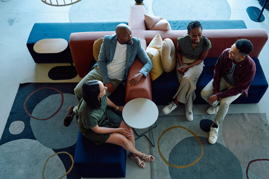 Group of diverse coworkers sitting on colorful sofas having casual conversation in office lounge
